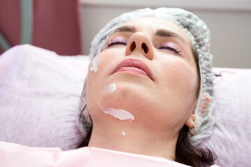 An adult brunette woman with her eyes closed after a facial rejuvenation procedure with cream on her face.