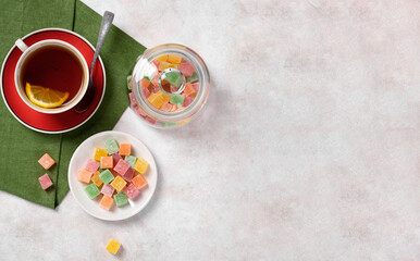 Obraz na płótnie Canvas Sweet candies in a jar. An overhead view of multi-colored marmalade cubes of Turkish delight on a white ceramic plate and in a jar on a light table and a cup of tea. banner with place for your text
