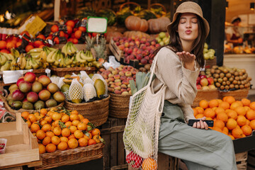Cute young caucasian woman closes her eyes and sends air kiss to camera sitting near market. Brunette wears hat, blouse, pants and string bag. Organic fruits concept