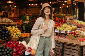 Nice caucasian young woman with shopping bag smiling looks at camera standing in vegetable row of market. Brunette wearing casual clothes is shopping on weekends. Lifestyle concept