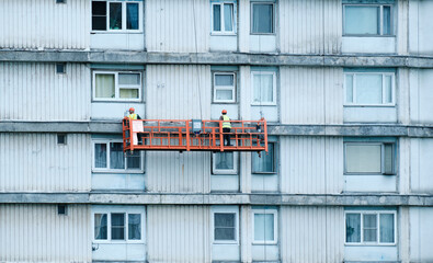 Two plasterers-painters are painting facade of house, standing in construction cradle. View of...