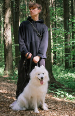 A young man stands with a white Samoyed dog on a leash in the forest and looks away. Walking with a dog in nature benefits the animal and the owner. Samoyed husky is a wonderful friend and companion.