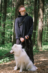 Young man stands with a white fluffy Samoyed dog in a summer forest and looks up. Walking with a dog in nature benefits the animal and the owner. The Samoyed husky is a wonderful friend and companion