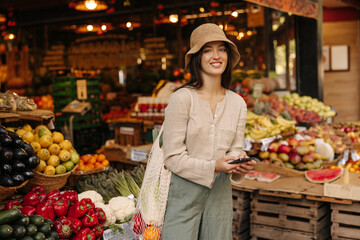 Cute young caucasian girl with smartphone looks at camera standing in market. Brunette wears casual clothes, hat and shopping bag. Lifestyle concept