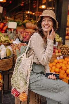 Cheerful young caucasian woman with shopping bag laughs looking away at vegetable market. Brunette wears casual clothes, hat. Healthy food concepts
