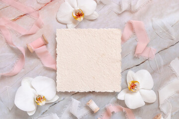 Handmade paper card near white orchid flowers and silk ribbons on marble, mockup