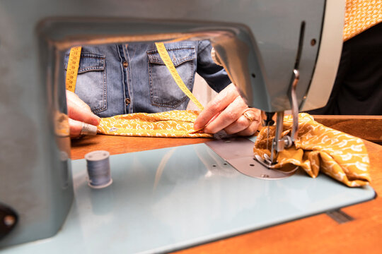 Hands Of Older Woman Measuring An Orange Fabric With A Tape Measure On The Wooden Furniture Of An Antique Sewing Machine