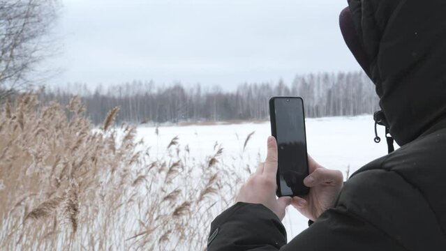 A man in a jacket and hood shoots a winter landscape on a phone camera. Shooting a snowy field, dry grass and forest