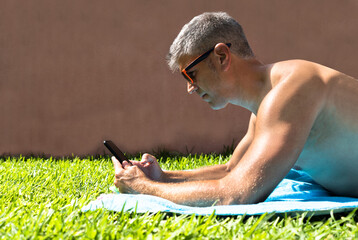 Man lying on the grass by the pool sunbathing while is using a phone. Summertime concept