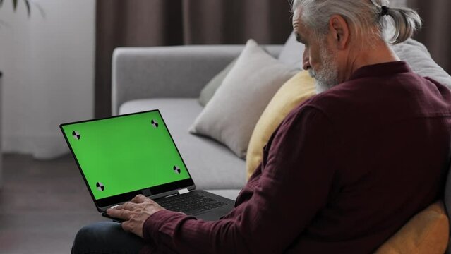 Elderly gray-haired man making video call at laptop green screen. Senior man talking and listening during video chat with friends or colleagues. Over shoulder monitor screen view.