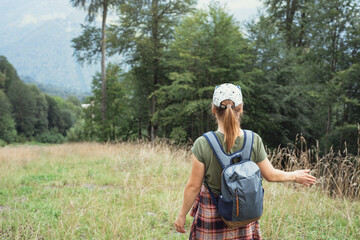 Naklejka na ściany i meble Rear view of a young woman in a cap with a backpack walking in the forest highlands in the summer and touching a growing plant enjoying nature
