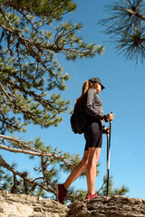 Young beautiful woman stay traveling with backpack on mountains with walking sticks on blue sky background and look away
