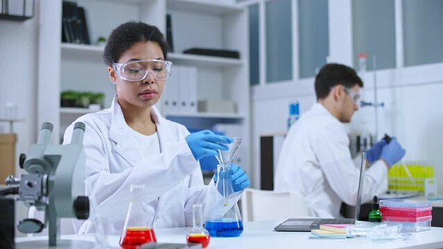 Female chemist mixing substances in lab, typing experiment report on laptop