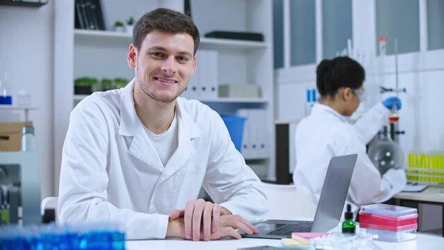 Professional male scientist working on laptop in laboratory, smiling on camera