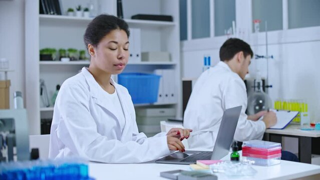 Black female scientist working on laptop in research lab, smiling on camera