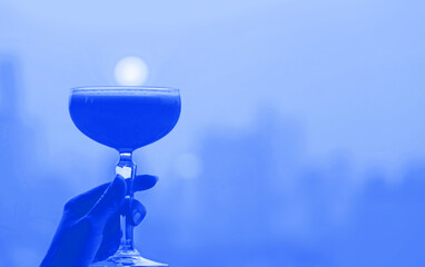 Pop art style cobalt blue colored cocktail glass in hand with blurry city aerial view in the backdrop