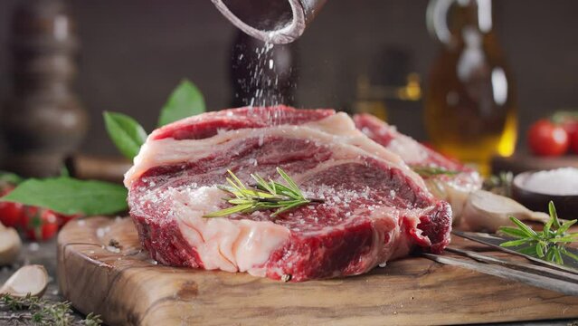 Two raw rib eye steaks with spices and herbs on a wooden kitchen table, sprinkle with sea salt on top. 