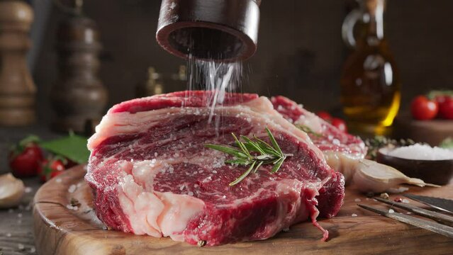 Two raw rib eye steaks with spices and herbs on a wooden kitchen table, sprinkle with sea salt on top. 