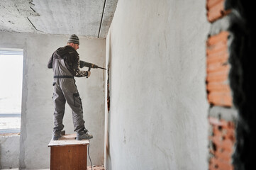 Full length of man builder standing on wooden desk and drilling wall with special construction tool. Male worker using electric drill machine while breaking the wall in apartment during renovation.