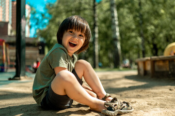 A three-year-old Asian boy with long black hair is sitting on the sand and wearing sandals. A child...