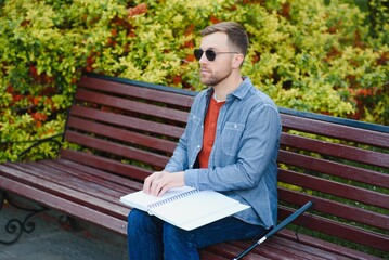 Fototapeta premium Blind man reading braille book, sitting on bench in summer park, resting