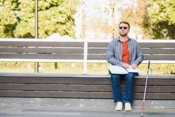 Visually impaired man with walking stick, sitting on bench in city park. Copy space