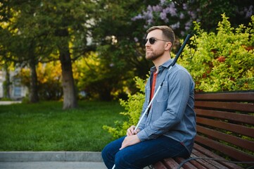Visually impaired man with walking stick, sitting on bench in city park. Copy space