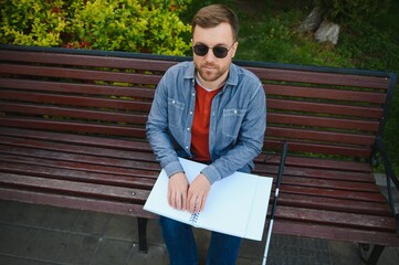 Fototapeta premium Blind man reading braille book, sitting on bench in summer park, resting