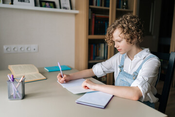 Medium shot of little child schoolgirl writing in exercise book doing some homework, sitting at table near window. Cute curly small school girl studying alone making notes at home.