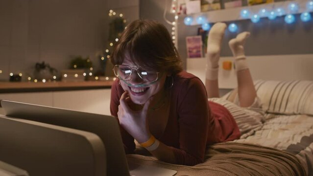 Full shot of young Caucasian woman with nasal cannula lying on bed in decorated hospital room, watching funny online videos on portable computer, laughing
