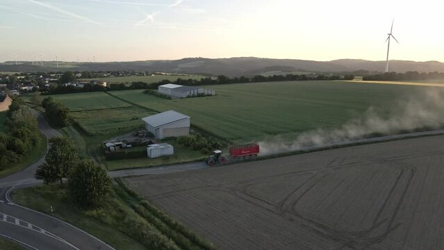 Dust trail forms from behind a large green tractor towing a red trailer through a green crop field. Drone shot.