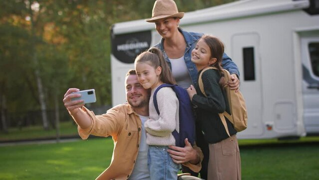 Happy young family with two children taking selfie with caravan at background outdoors.
