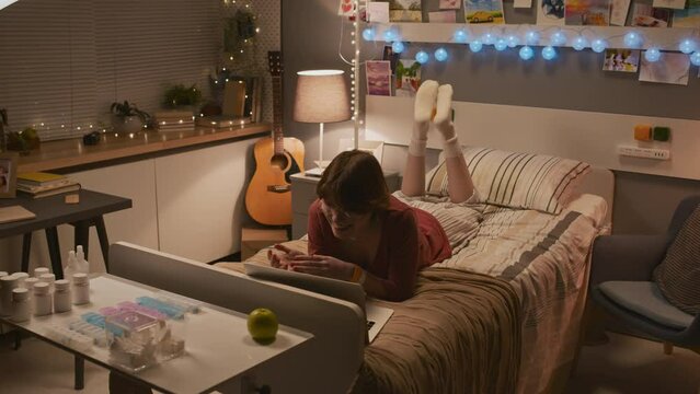 Wide high angle of young Caucasian woman with nasal cannula lying on bed in decorated hospital room, watching online videos on laptop computer