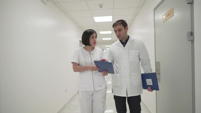 Two doctors man and woman, discussing case reports and medical documents as they walk down corridor in hospital. Chief doctor and general practitioner discuss reports on clipboard in clinic challway