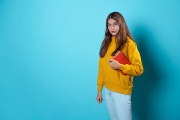 A young student looks at a book and reads. Education and training, visiting the library. Young woman in yellow hoodie and jeans on a blue background, copy space