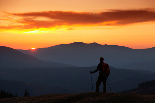 A man with a backpack is outdoor at sunset. Orange sky. Silhouettes of mountains peaks