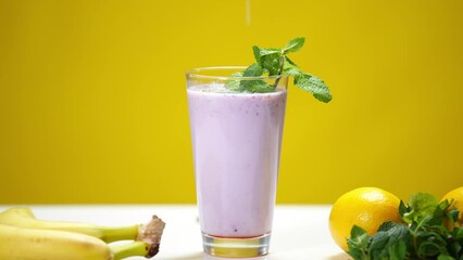 Close-up purple vitamin smoothie with mint and lemon juice drops falling in slow motion in drink. Refreshing organic dairy and fruit cocktail at yellow background