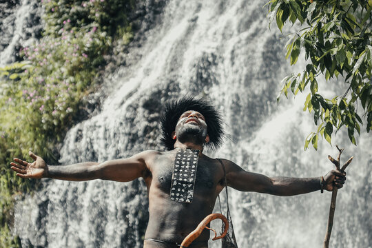 Papua man of Dani tribe spread out his hands feeling freedom, against waterfall at greenery forest.