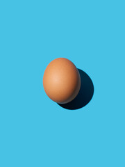 A brown chicken egg lies in the center on a bright blue background. Bright light, deep shadow, vertical. Close-up, copy space, top view.