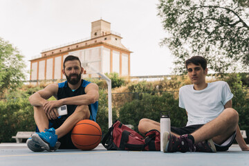 Fototapeta premium Two young boys sitting resting after a street basketball practice holding a smartphone. Partnership concept.