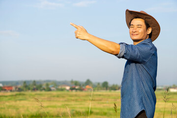 Portrait of handsome Asian man farmer wears hat, blue shirt, points to  sky background. Copy space for adding text or advertisement. Concept : Agriculture occupation. Happy farmer.