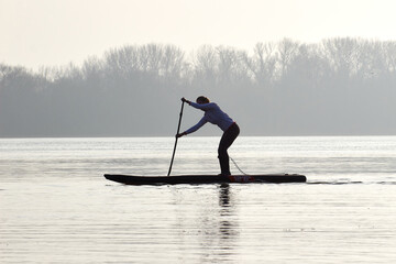 Silhouette of sporty woman rowing on stand up paddle (SUP) at calm winter Danube river against the background of the shore with trees