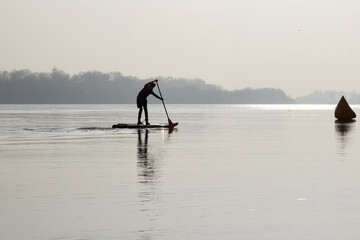 Fototapeta na wymiar Silhouette of a boy paddle on stand up paddle boarding (SUP) on quiet autumn river at the morning. Morning training and meditation on the water