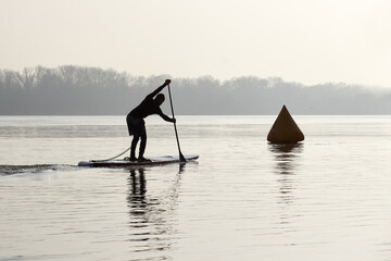 Fototapeta na wymiar Silhouette of a boy paddle on stand up paddle boarding (SUP) near buoy on quiet autumn river at the morning. Morning training and meditation on the water