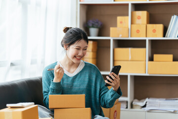 Happy young asian woman startup small business freelance holding parcel box and computer laptop and sitting on sofa, Online marketing packing box delivery concept.