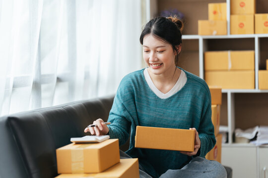 Happy Young Asian Woman Startup Small Business Freelance Holding Parcel Box And Computer Laptop And Sitting On Sofa, Online Marketing Packing Box Delivery Concept.