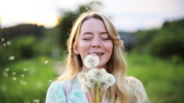 beautiful young blonde woman with long hair blowing on a dandelion in a field at sunset, fabulous video in nature, summer vacation concept.
