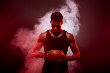 Basketball player side lit with red color holding a ball against smoke background. Serious...