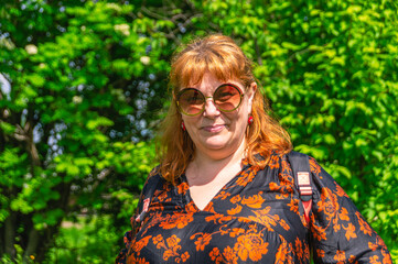 A plump red-haired woman in round sunglasses with a smile against a background of green trees. A woman in an orange dress in the countryside. Positive mood in nature. Joy on a sunny day.