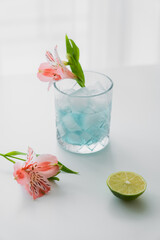 glass with iced tonic water near pink alstroemeria flowers and lime on white surface.